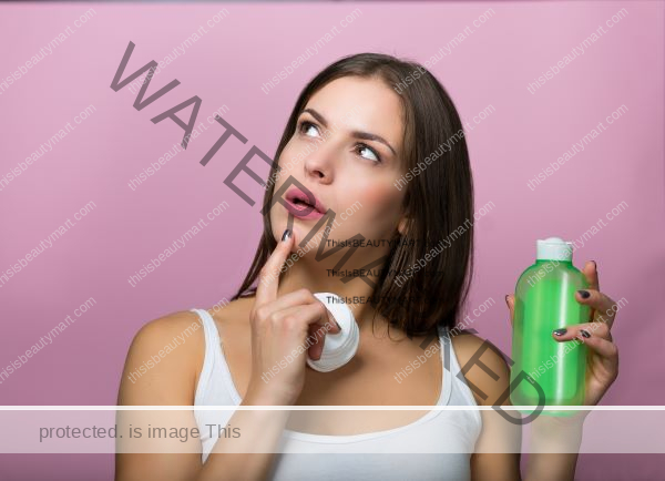 A woman holding a bottle of a generic cosmetic product and some applicators, while wondering about something