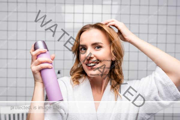 A woman holding a bottle of hair care product and applying it to her hair in a bathroom, wearing a bath robe