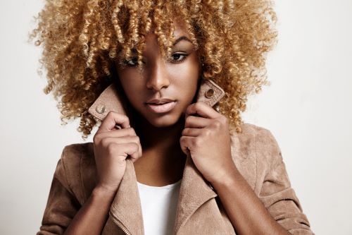 Black woman with curly hair dyed blonde, posing fashionably with a brown jacket