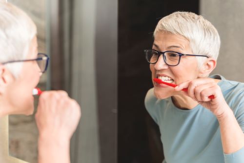 Gray-haired senior-looking woman brushing her teeth in front of her bathroom mirror